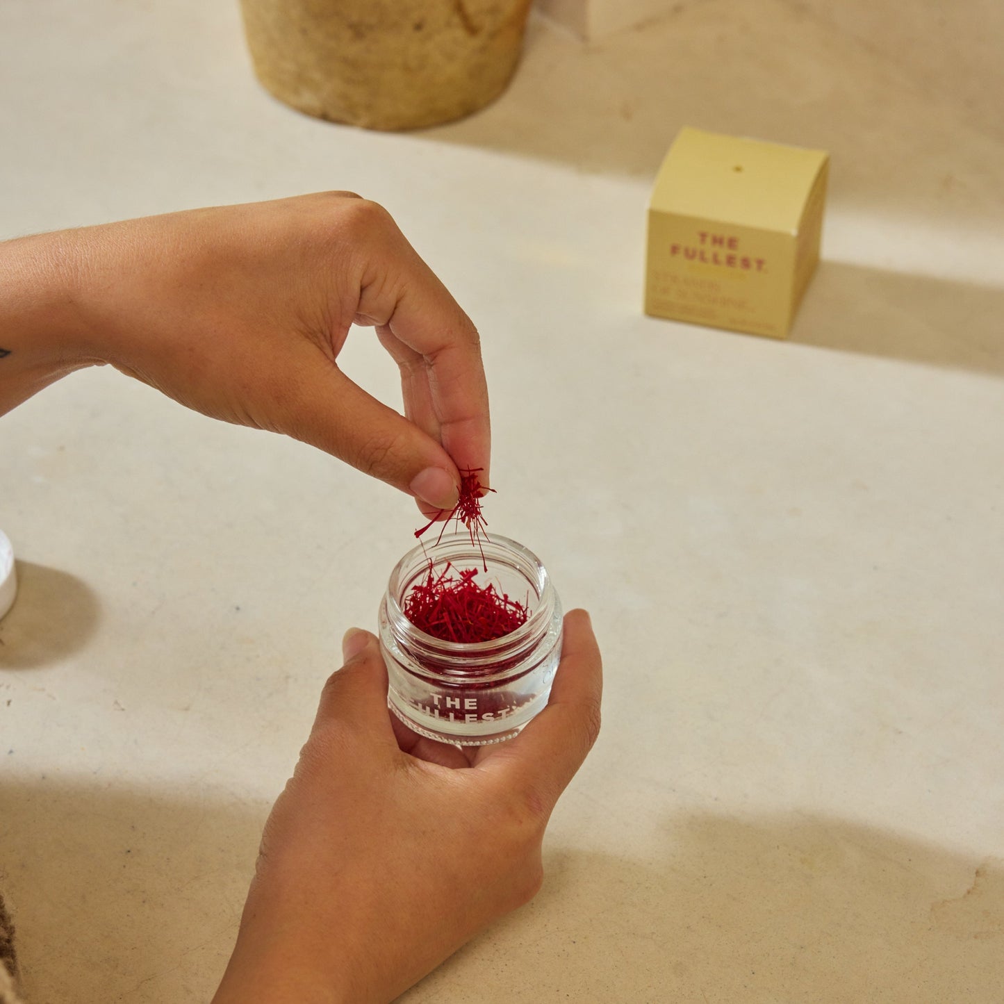Hands place Saffron Threads into a glass jar on a light countertop, with a yellow box labeled "THE FULLEST" in the background, highlighting this premium product.